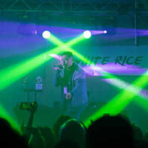 Artist HMisfits performing on stage holding a microphone, audience in front of him with green stage lights beaming into crowd. HMisfits is an Asian artist that won an award in Chippewa Valley.