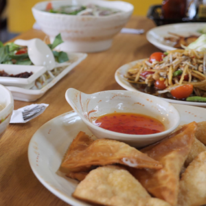 A photo of the food available at Thai Orchid restaurant. In the image on white plates are crag rangoons with sweet and sour sauce in a cup. Behind it is papaya salad and other foods.