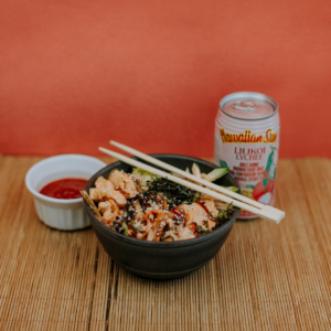 Picture of a Hawaii Poke Bowl with chopsticks balancing on top. On the side is a red condiment in a white cup. To the right is a Hawaiian Sun Lychee can drink.